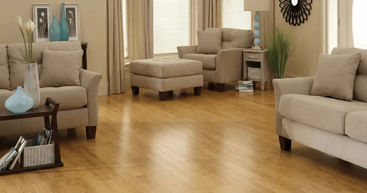 A living room with professionally installed bamboo flooring, beige sofas, a matching ottoman, a side table with a blue lamp, vases, framed pictures, and large windows with blinds and curtains in Anaheim, CA