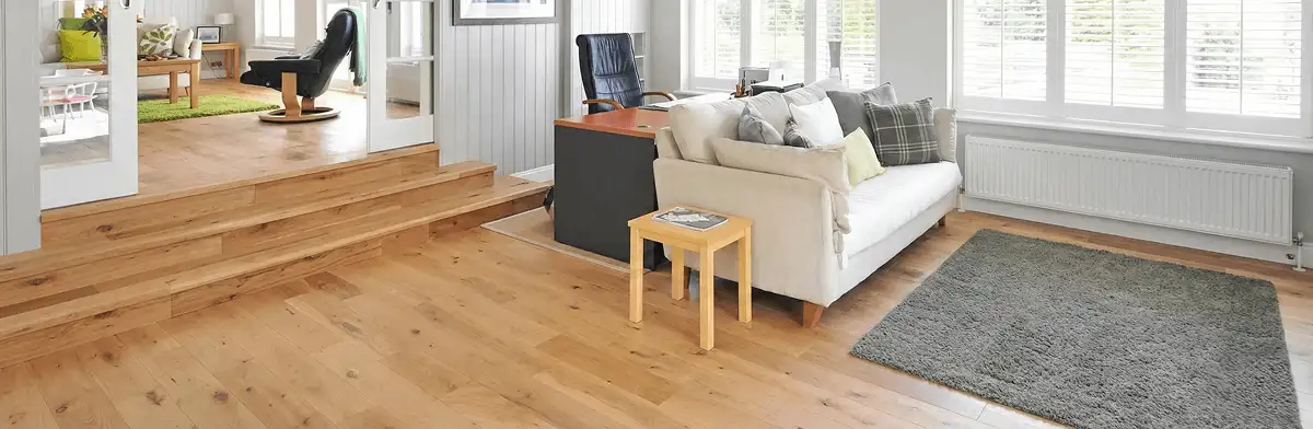 A modern living room with professionally installed laminate flooring, a beige sofa, a small side table, a gray rug, and large windows with white shutters in Anaheim, CA